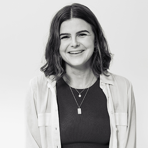 A smiling woman with shoulder-length wavy hair, wearing a dark top and light jacket, standing against a plain background.