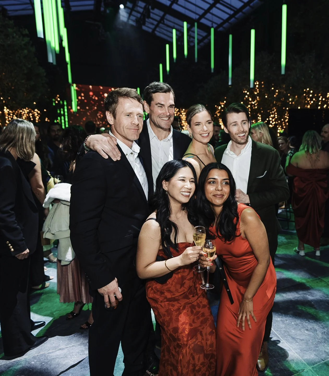 A group of six people at a celebration, holding glasses of champagne, dressed in formal attire, smiling at a party with green and yellow lights and a festive atmosphere.