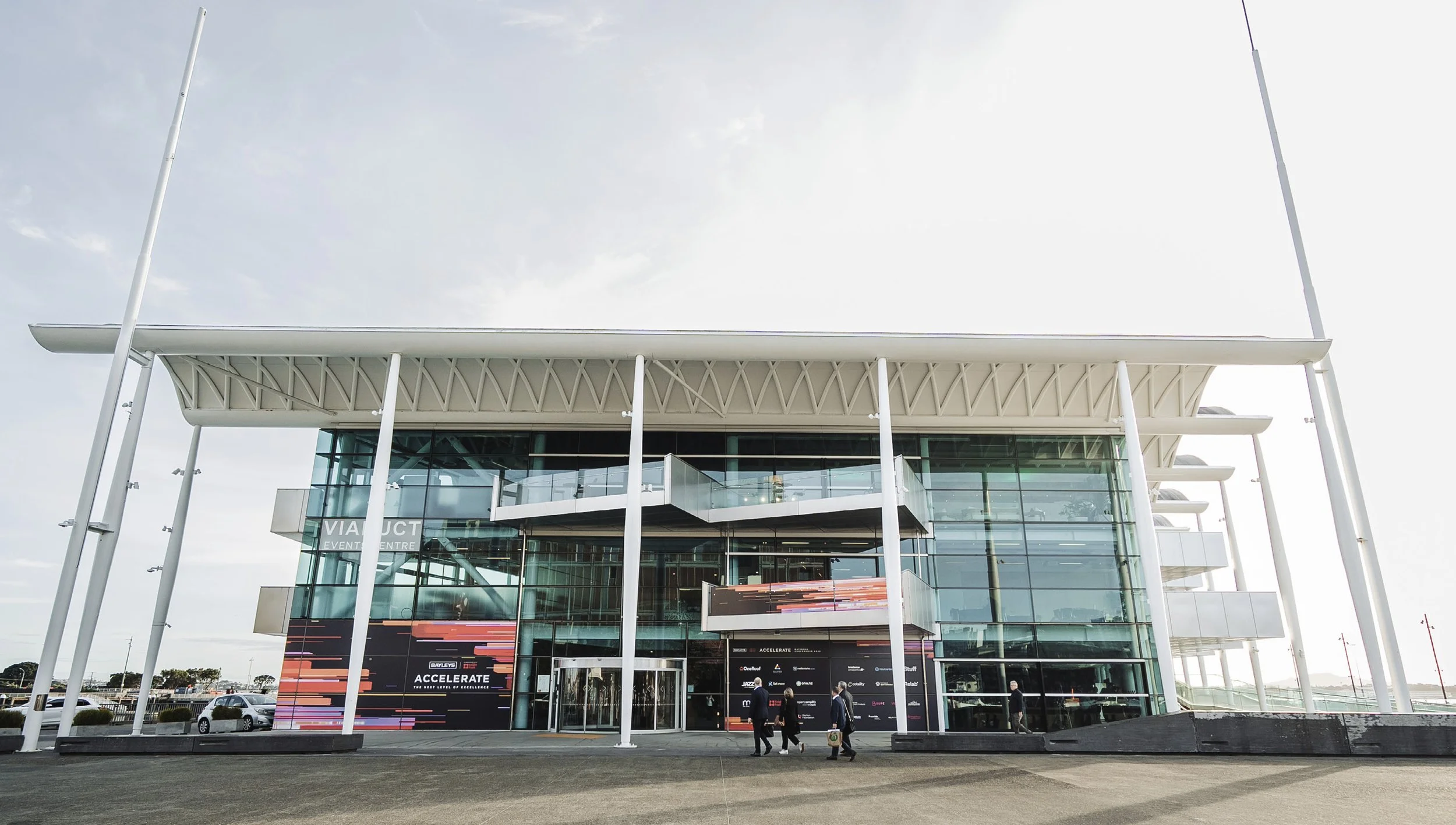 Modern building with glass facade and white structural supports, sign reading 'VIADUCT EVENT CENTRE,' people walking in front, and a digital display board near the entrance.
