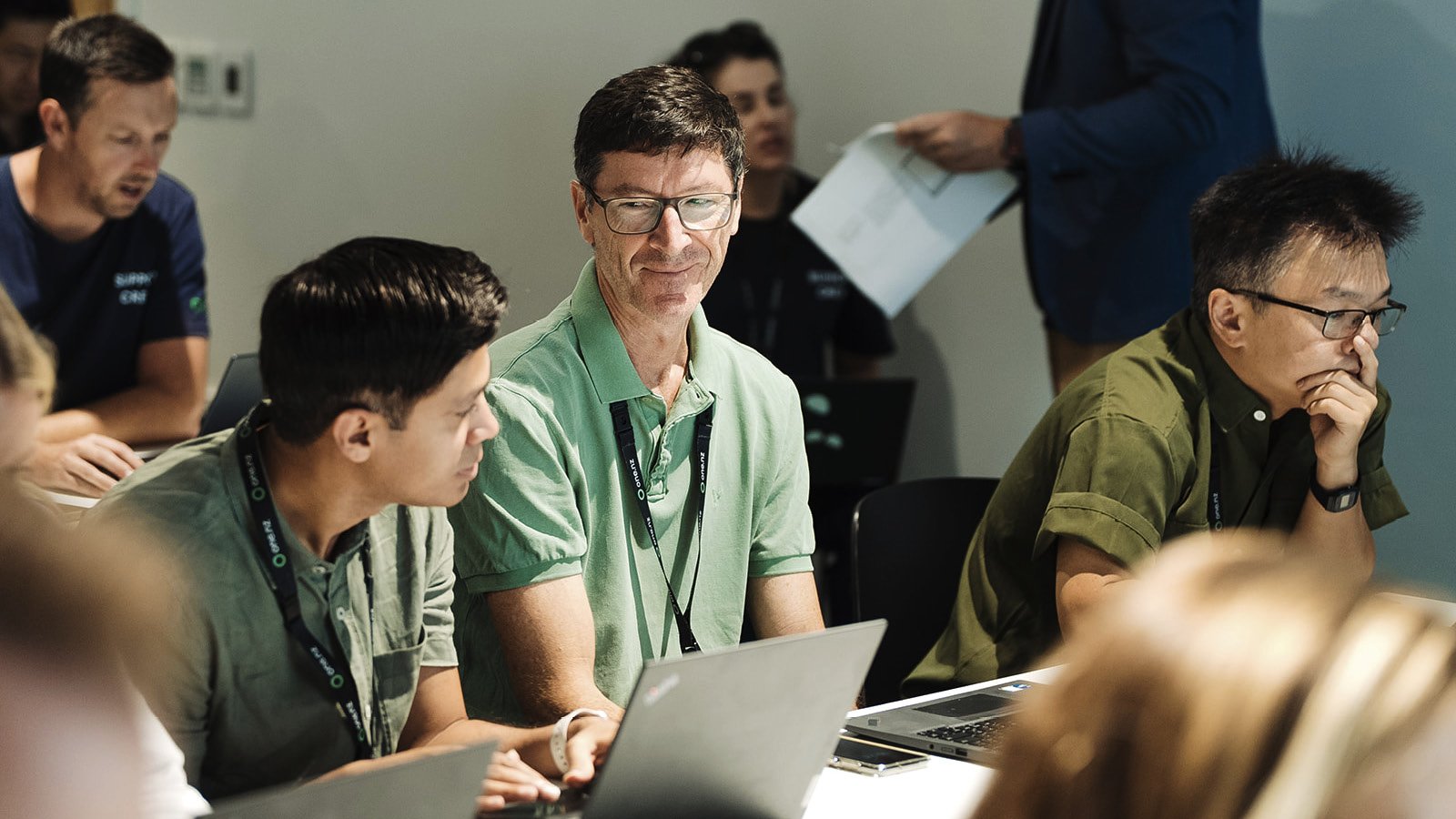 A man with glasses and a green polo shirt looks at a laptop during a meeting, surrounded by other professionals with laptops, in a conference room.