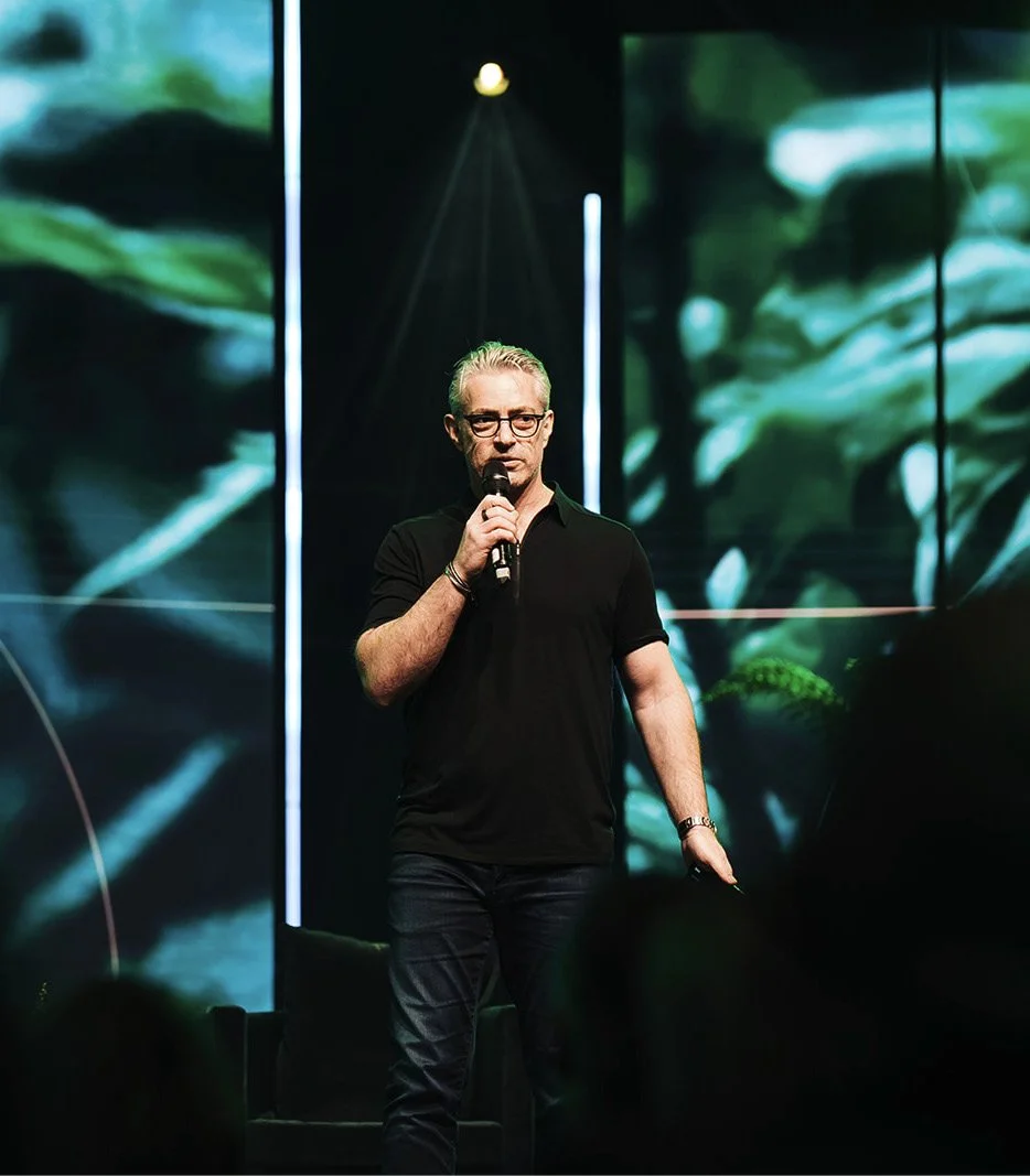 A man with glasses and gray hair is standing on a stage, holding a microphone, with a dark background and some abstract visuals or screens behind him.