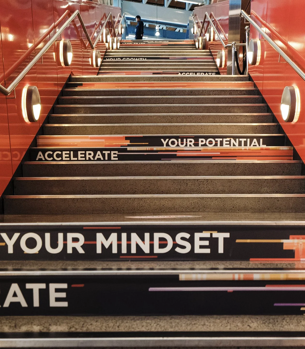 Staircase with motivational messages on each step, including 'Your Mindset,' 'Accelerate,' 'Your Potential,' and 'Your Growth.' A person is at the top of the stairs, in an indoor space with people and glass walls.