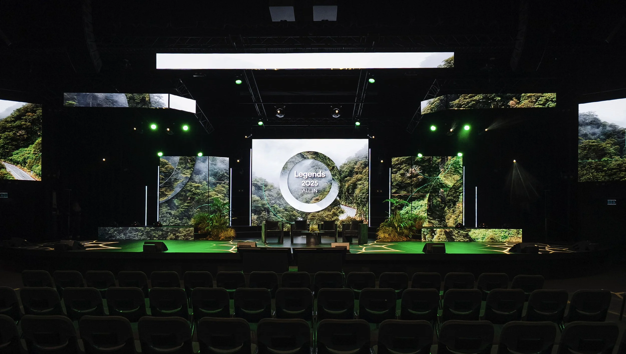 An empty stage set up for a conference with a large central screen displaying "Legends 2025 ALL IN" and scenic nature visuals, surrounded by smaller screens and green stage lighting, with chairs facing the stage.