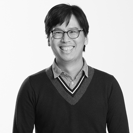 Black-and-white photo of a smiling man with glasses wearing a collared shirt and a sweater with a V-neck.
