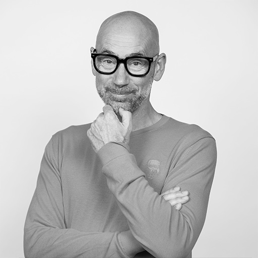 Black and white portrait of a bald man with glasses, wearing a long-sleeve shirt, posing with his hand on his chin and crossed arms.