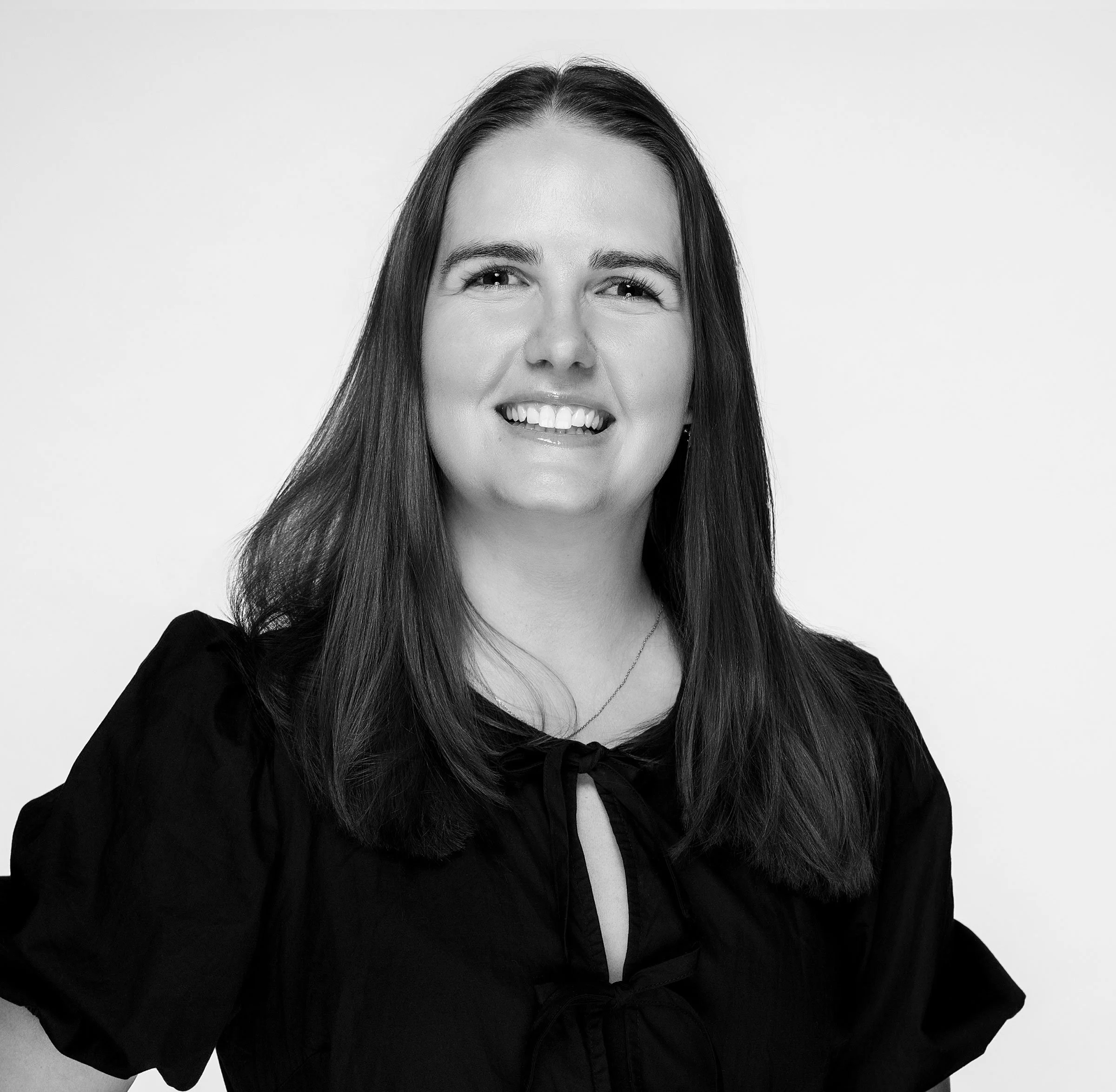 Black and white photo of a woman with long hair, smiling, wearing a black top and a necklace, against a plain background.