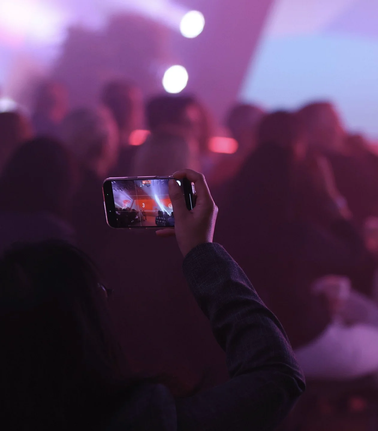 Person taking a photo or video of a stage with their smartphone at an indoor event or concert, with a blurred audience and pink and purple lighting.