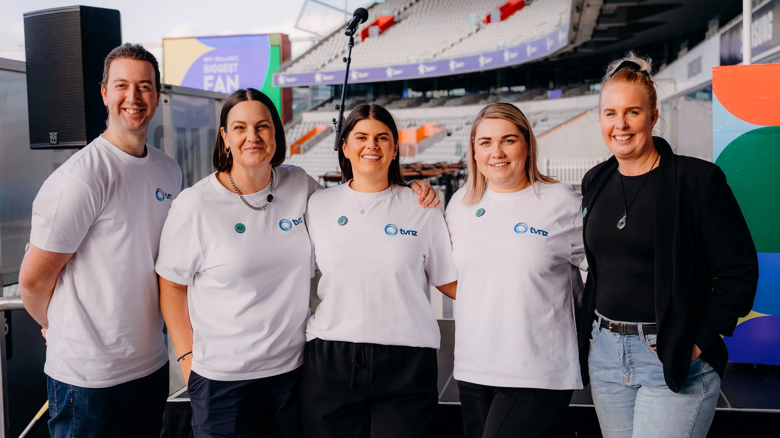 Group of five people smiling at an outdoor event at a stadium, wearing white t-shirts with the TVNZ logo, standing in front of a large screen and colorful background.