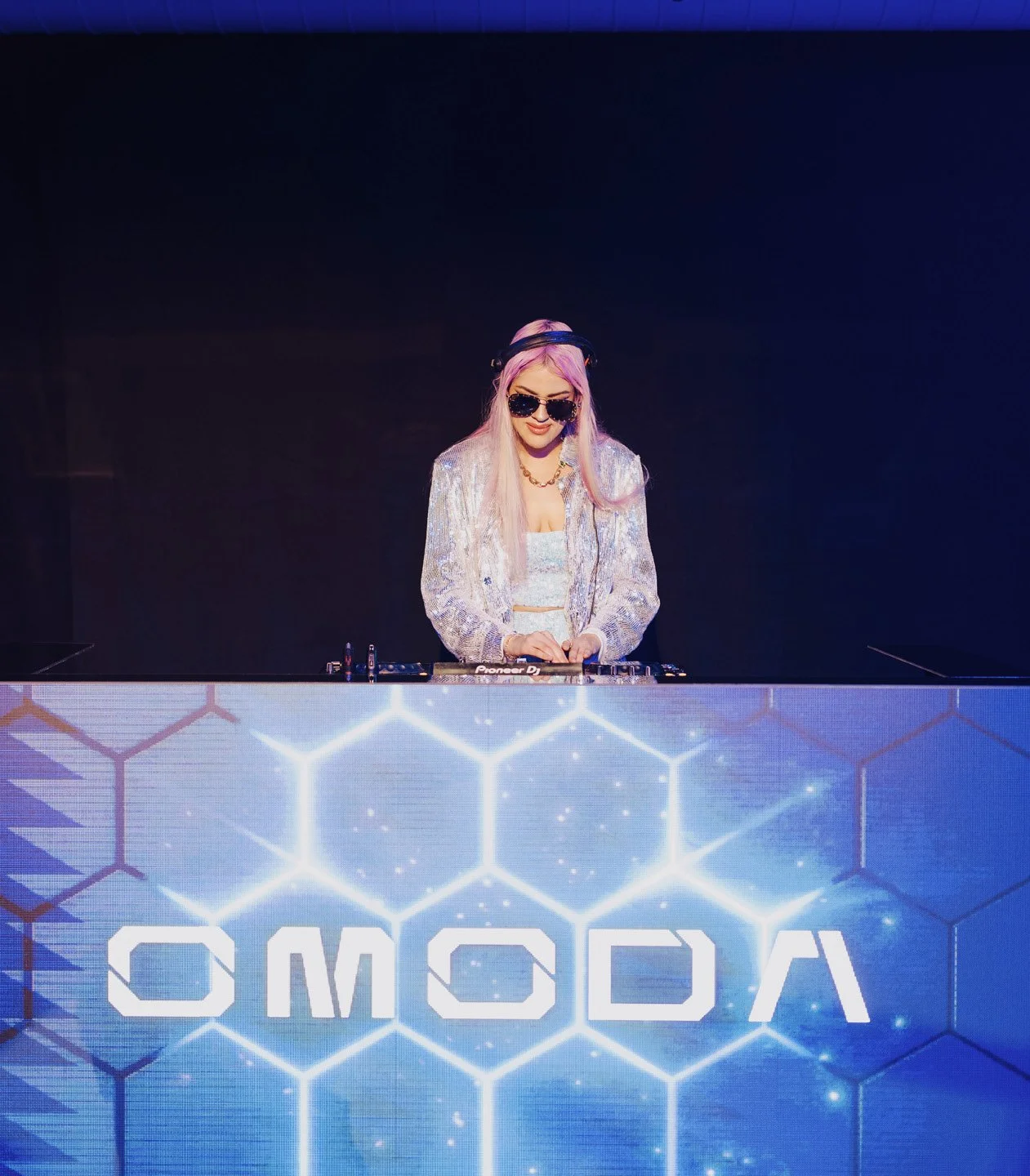 Female DJ with pink hair, sunglasses, and headphones, performing at a club, illuminated by blue and white hexagonal graphics.