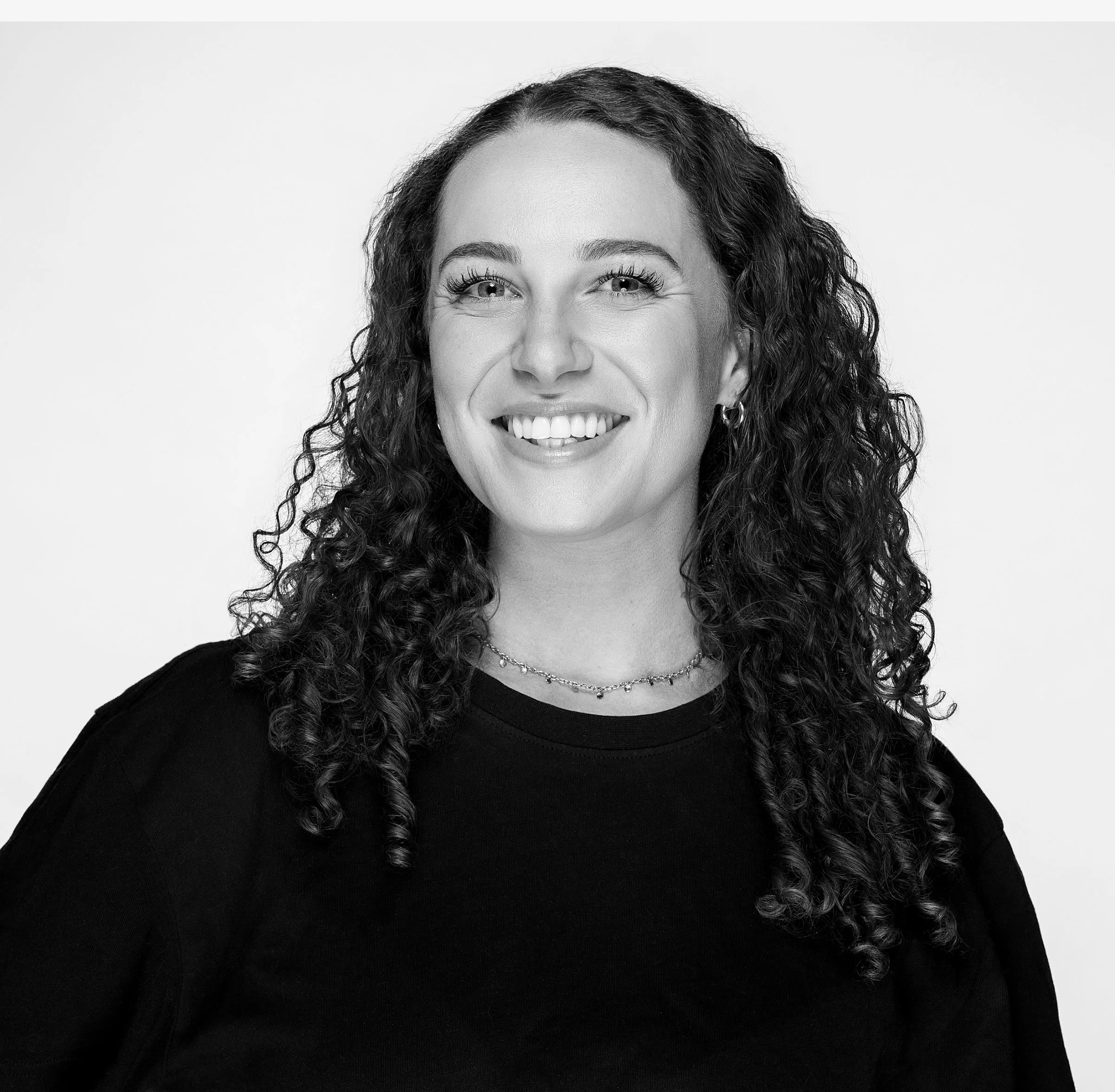 Black and white portrait of a smiling woman with curly hair, wearing a light-colored t-shirt and a dark sleeveless top.