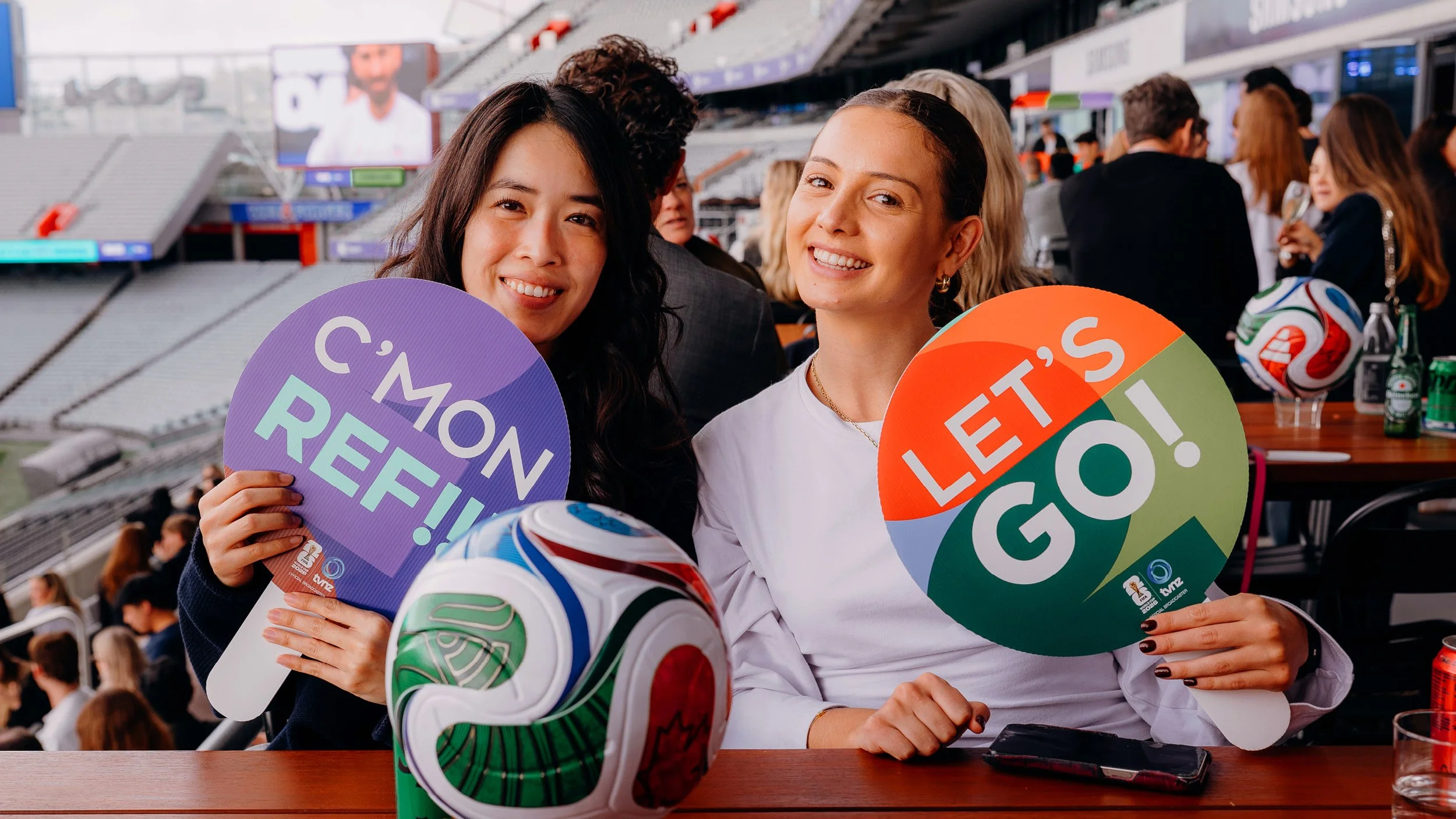 Two smiling women holding colorful round signs that read 'C'MON REF'! ''LET'S GO!' at a sports stadium, with a crowd in the background and a soccer ball on the table in front of them.