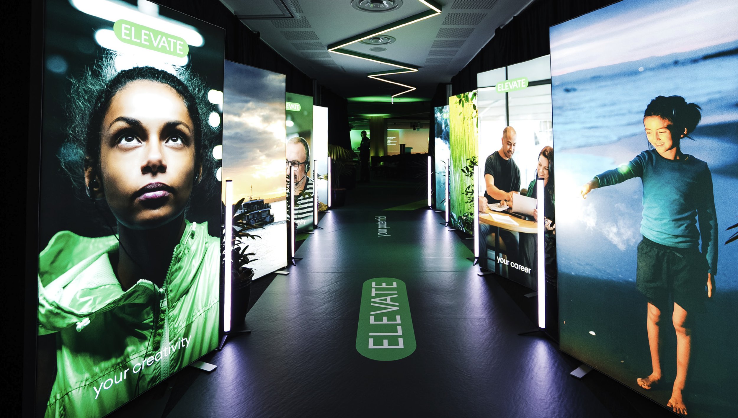 A hallway with illuminated posters featuring diverse people, with the word "ELEVATE" on the posters and floor, promoting creativity and career growth.