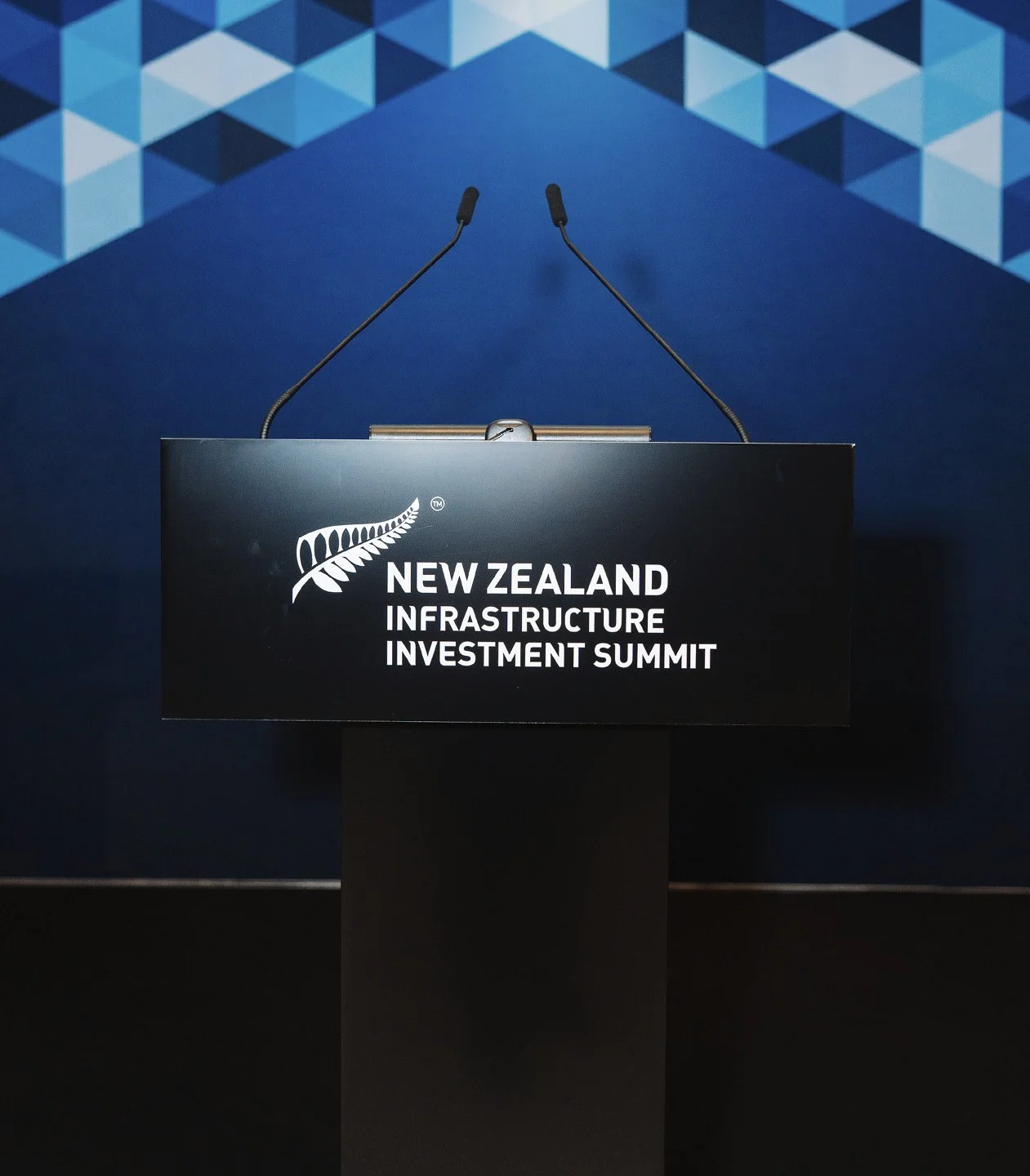 A black podium with two microphones and the logo for the New Zealand Infrastructure Investment Summit in front of a blue and white geometric patterned backdrop.
