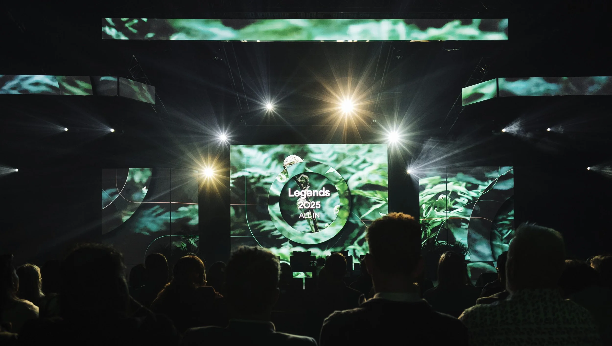 A large conference or presentation event with an audience facing a stage showing a digital screen. The screen displays "Legends 2025 ALL IN" with a green nature-themed background. Bright stage lights illuminate the scene.