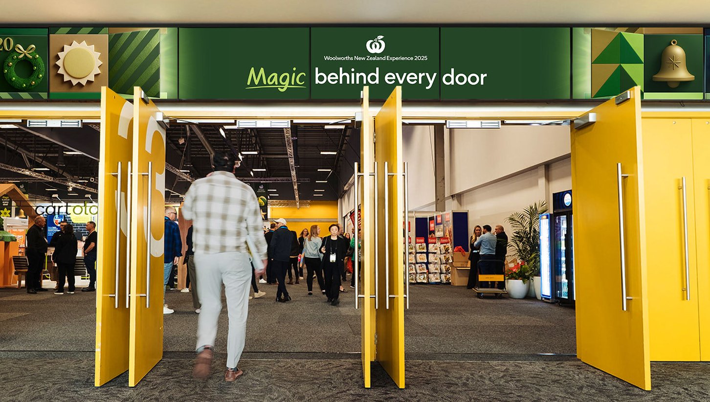 People walking into a trade show inside a large hall with yellow doors, green signage that reads 'Magic behind every door', and various booths and displays.