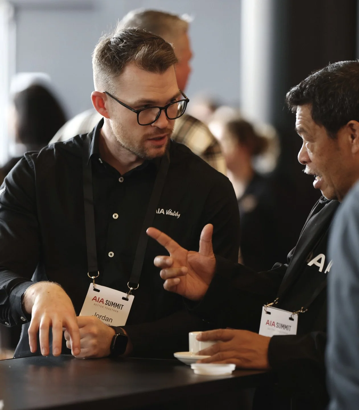 Two men in conversation at an event, one with glasses and a watch, the other with a cup, wearing name tags labeled 'AIA Summit'.