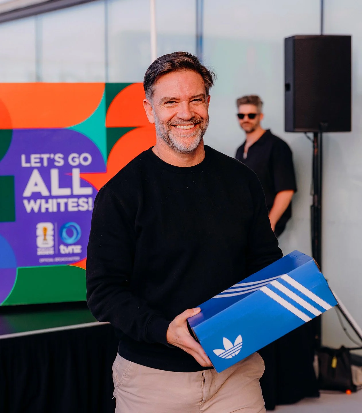 Smiling man holding a blue Adidas shoe box at an event, with a colorful sign in the background that says 'Let's go all whites!'