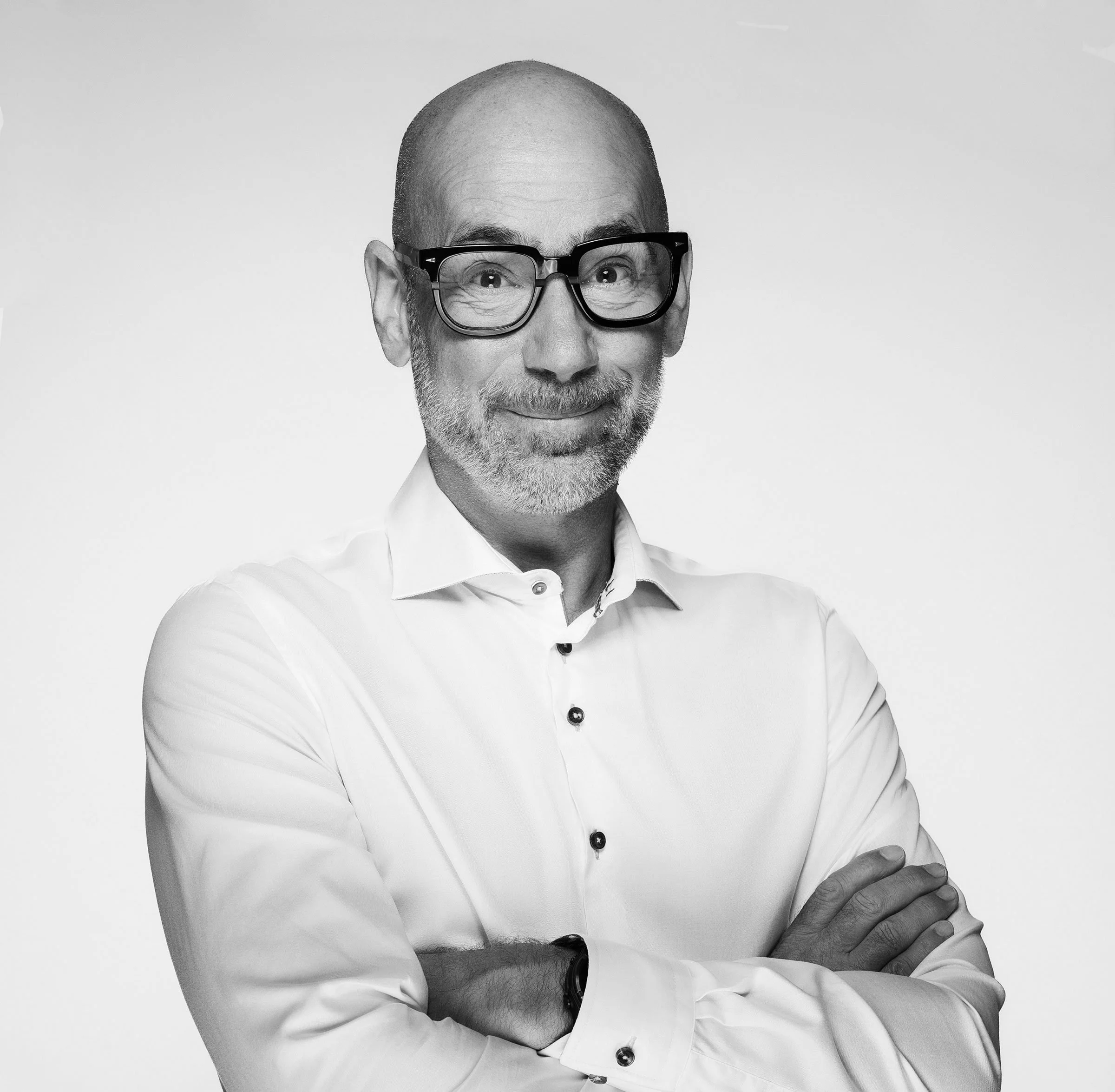 Black and white portrait of a bald man with glasses, wearing a long-sleeve shirt, posing with his hand on his chin and crossed arms.