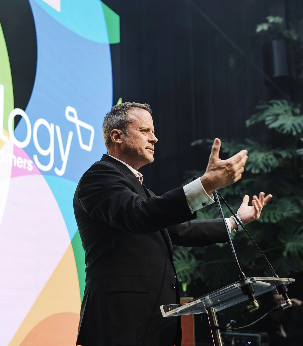 A man in a black suit giving a speech at a podium, with large colorful branding and green plants in the background.