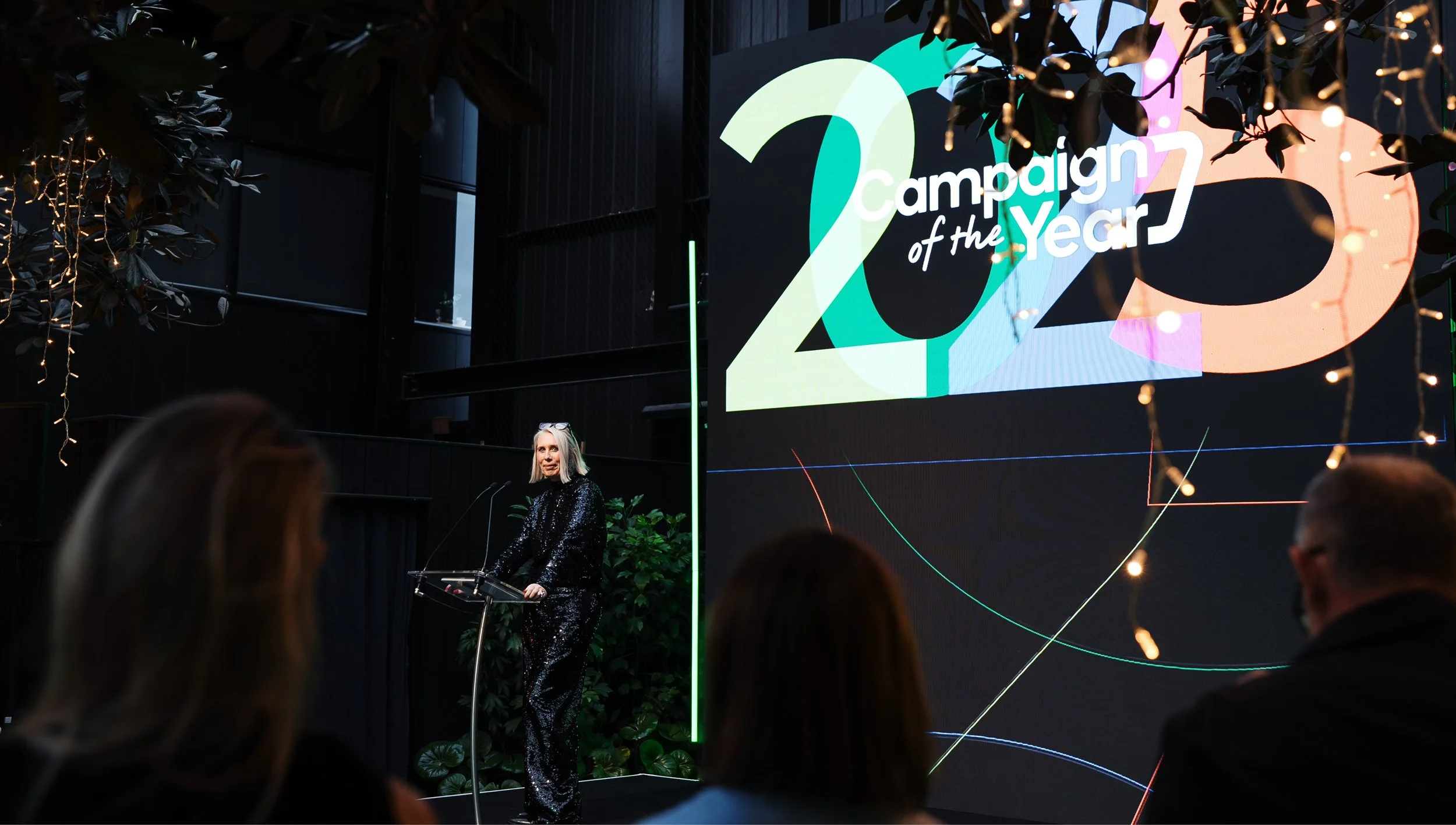 A woman stands at a podium during an event titled 'Campaign of the Year 2023,' with a large screen displaying the event name in the background, decorated with string lights and foliage.