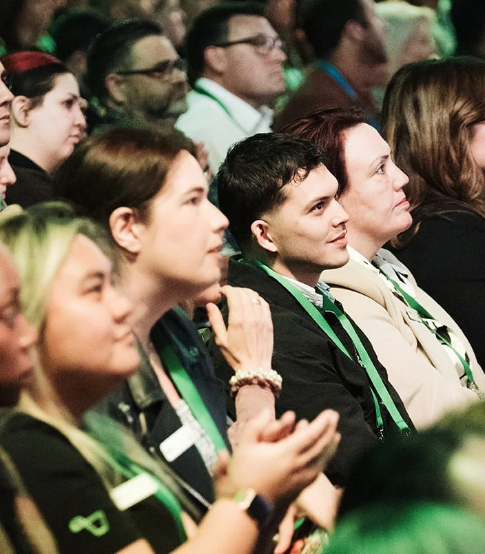 Audience members attending a conference or event, seated closely, wearing ID badges with green lanyards, attentively listening.