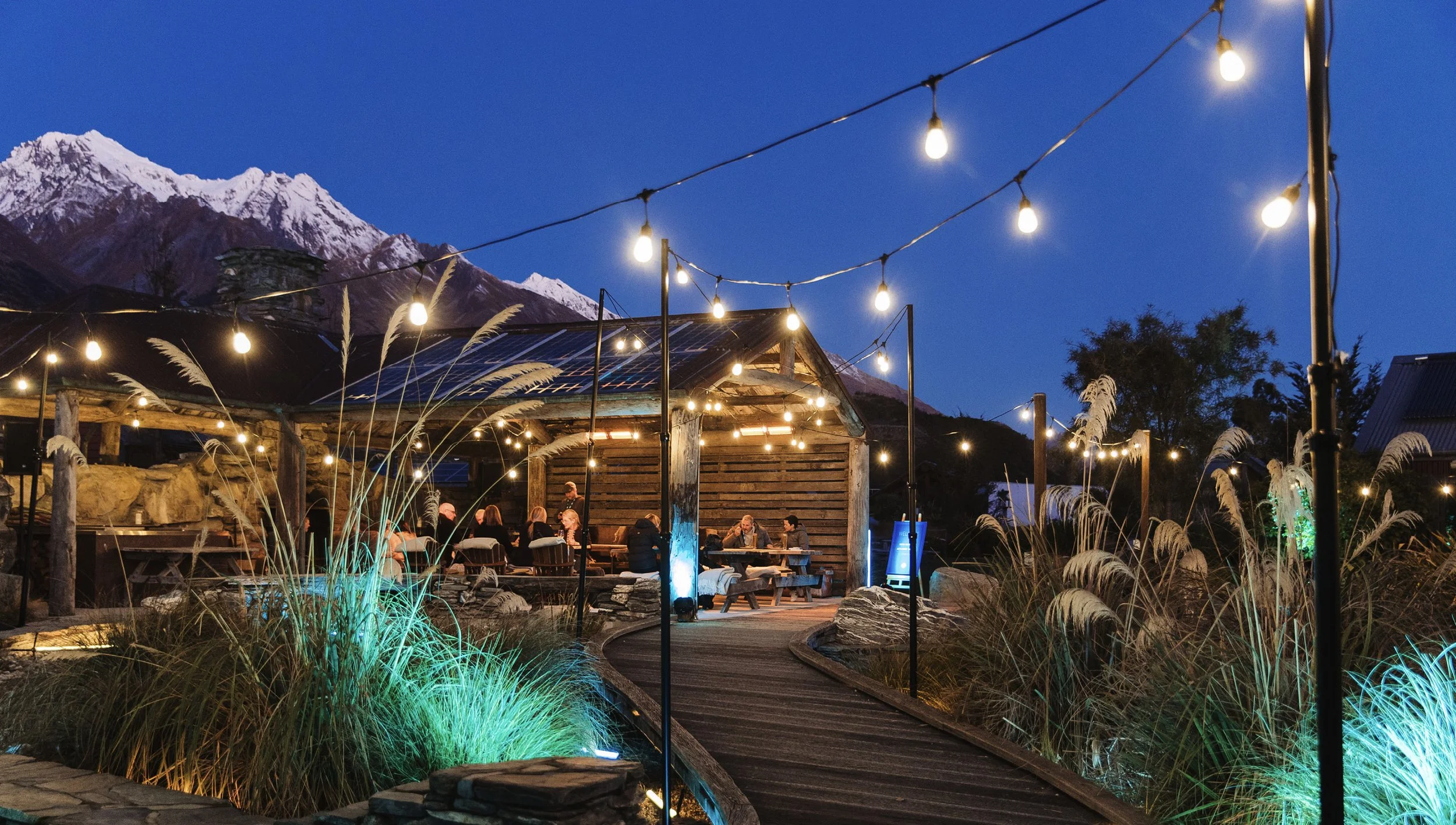 Outdoor restaurant or bar area at night with string lights, seating, and a mountain range with snow-capped peaks in the background.
