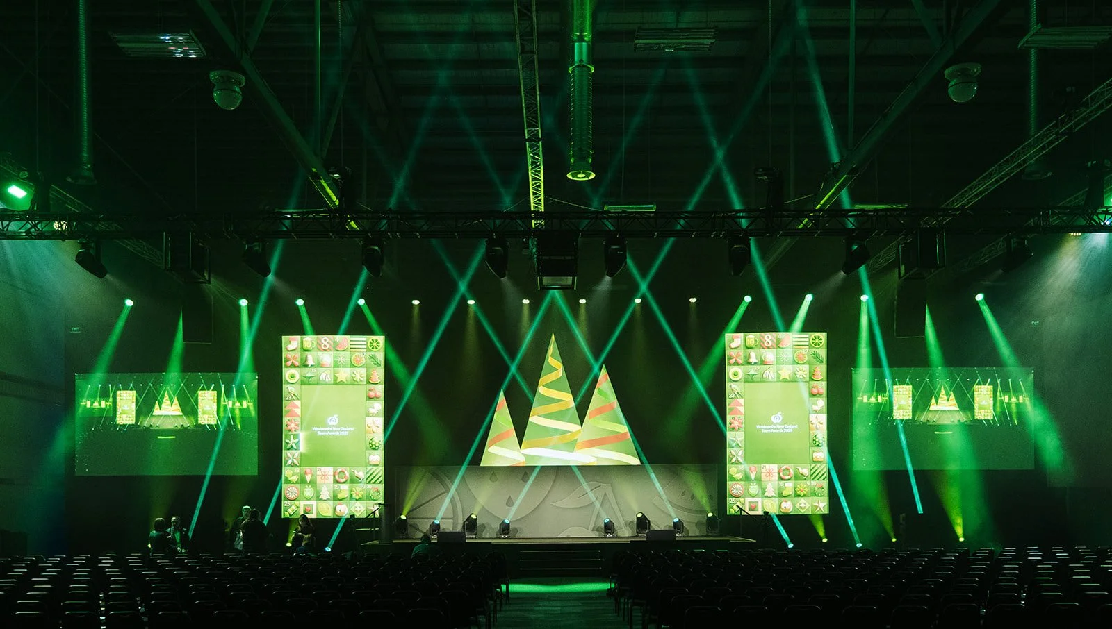 Empty event stage with green lighting and holiday-themed decorations, including Christmas trees and colorful digital screens.
