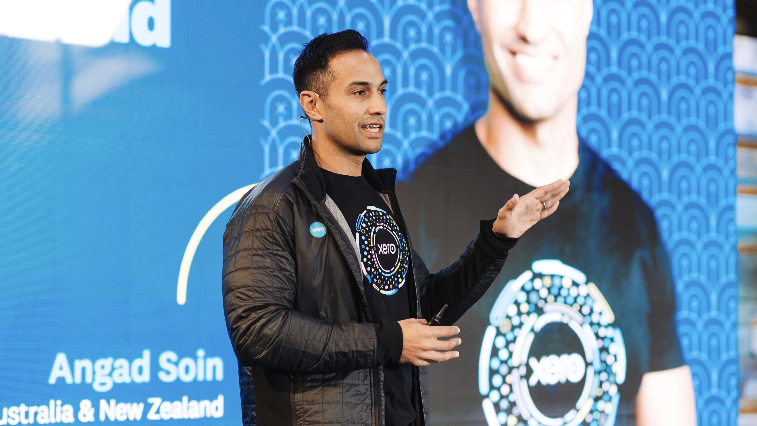 Man speaking at a conference or event, standing in front of a large screen with graphics and text that mentions Angad Soin from Australia and New Zealand, with the Xero logo on his shirt and badge.