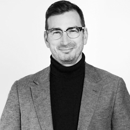 A man with glasses, wearing a suit jacket and turtleneck, smiling in front of a plain background.