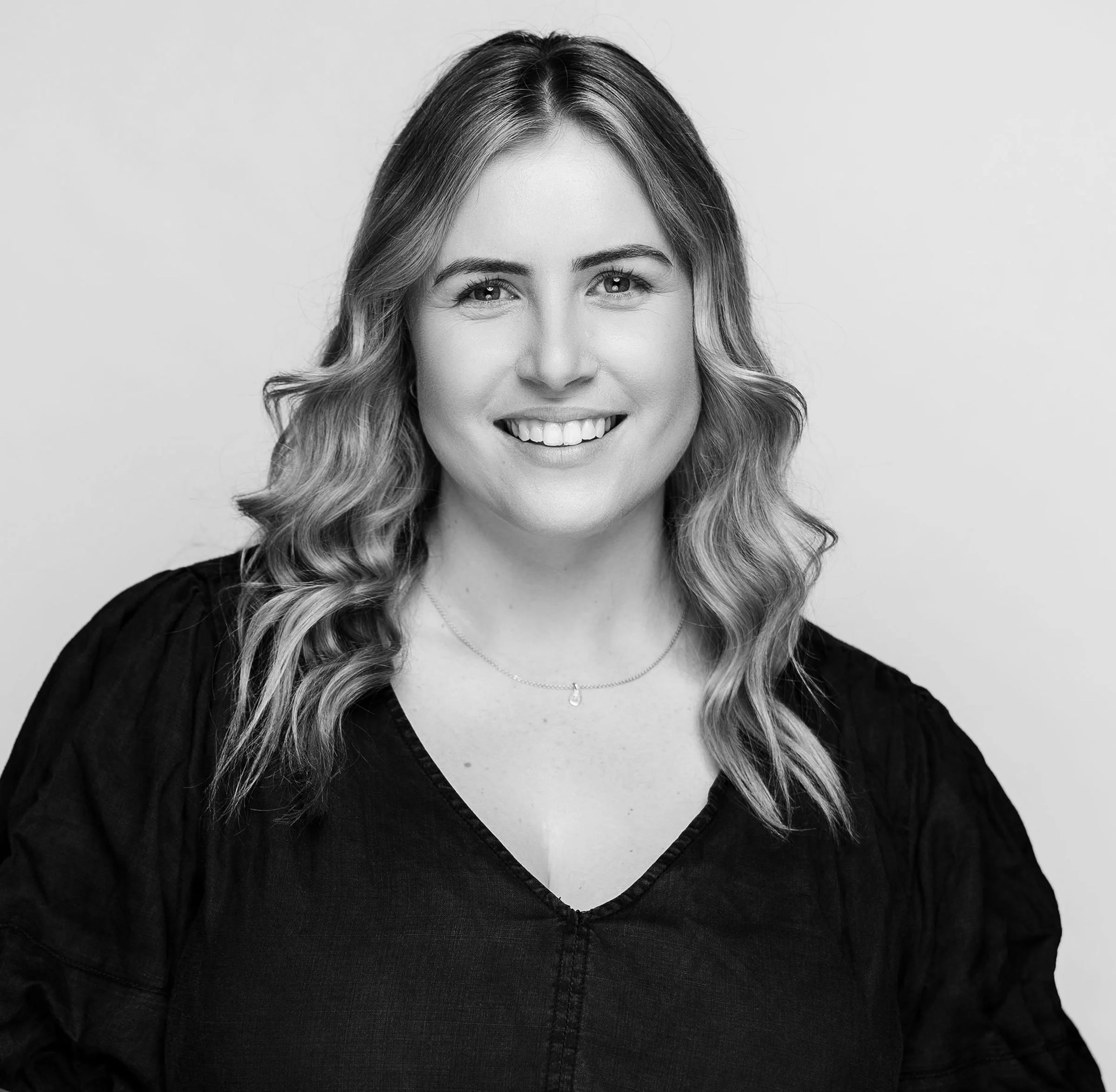Black and white photo of a smiling young woman with shoulder-length blonde hair, wearing a dark top, against a plain white background.