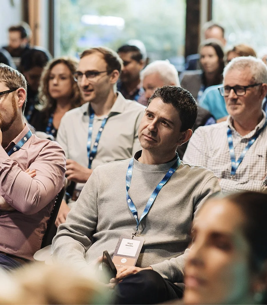 A group of people attending an indoor conference or seminar, listening attentively, with some wearing name badges and conference lanyards.