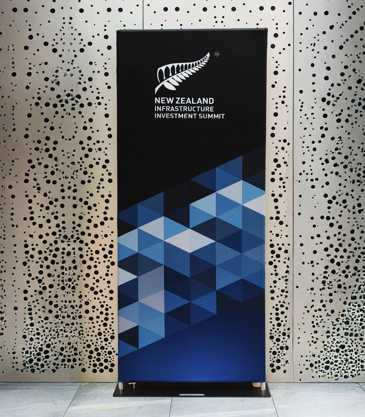 Black vertical banner with a silver fern logo at the top, displaying white text that reads 'New Zealand Infrastructure Investment Summit,' against a geometric blue patterned background, standing in front of a decorative perforated wall.