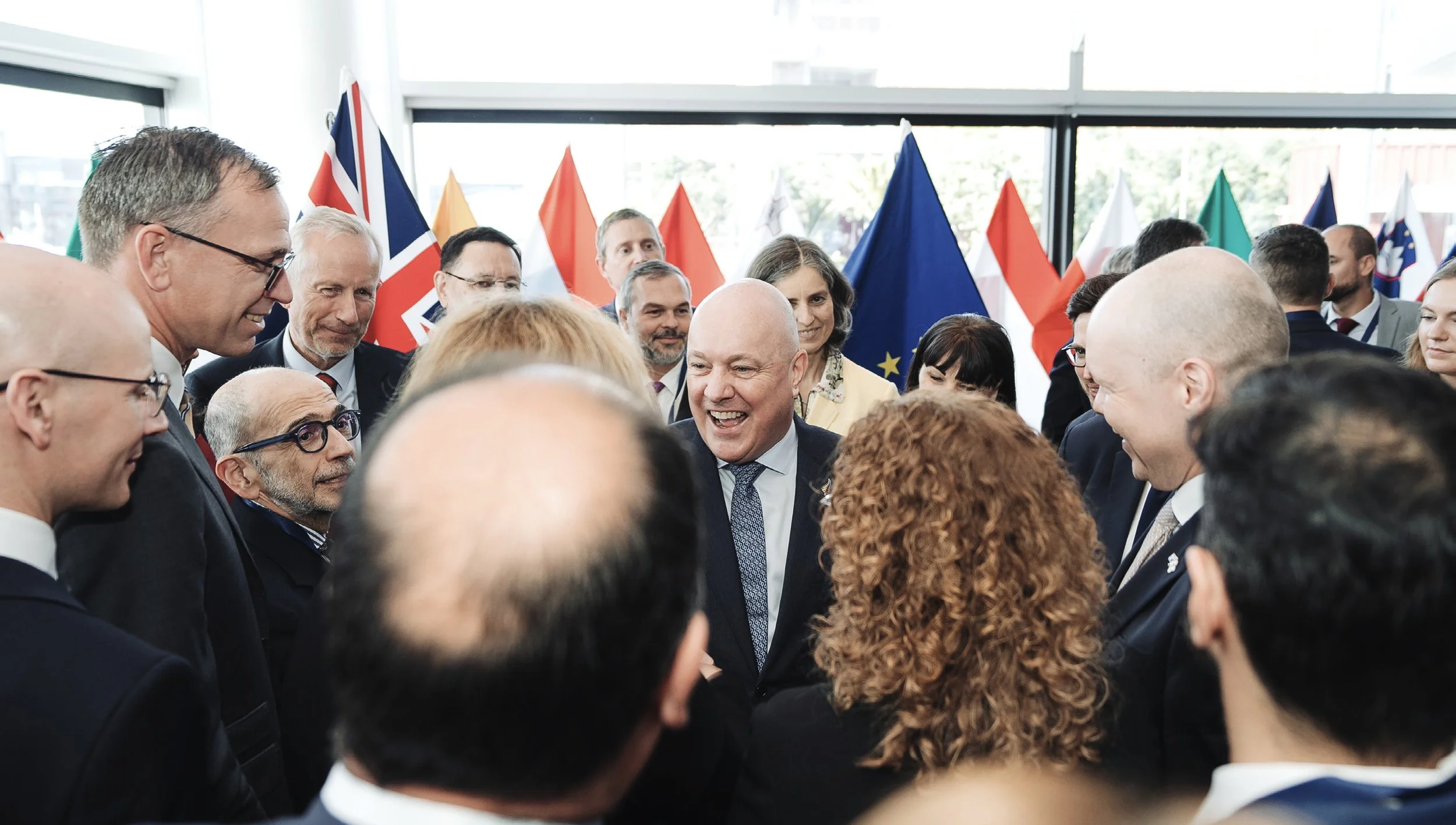 A group of people, mostly men in suits, standing in a circle and smiling or talking at an indoor event with flags of different countries in the background.