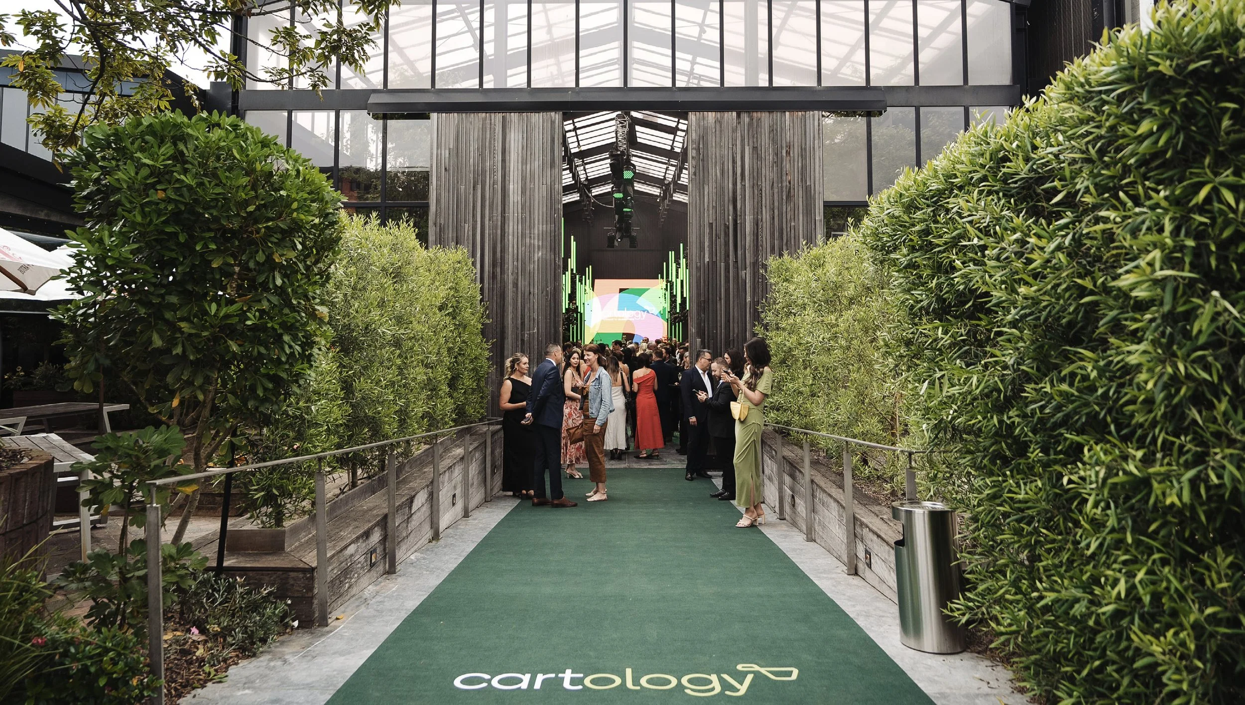 People gathered at an outdoor event on a green carpet entrance with plants on both sides leading to a modern building with large glass windows. There is a digital screen in the background displaying the logo and name 'Cartology'.