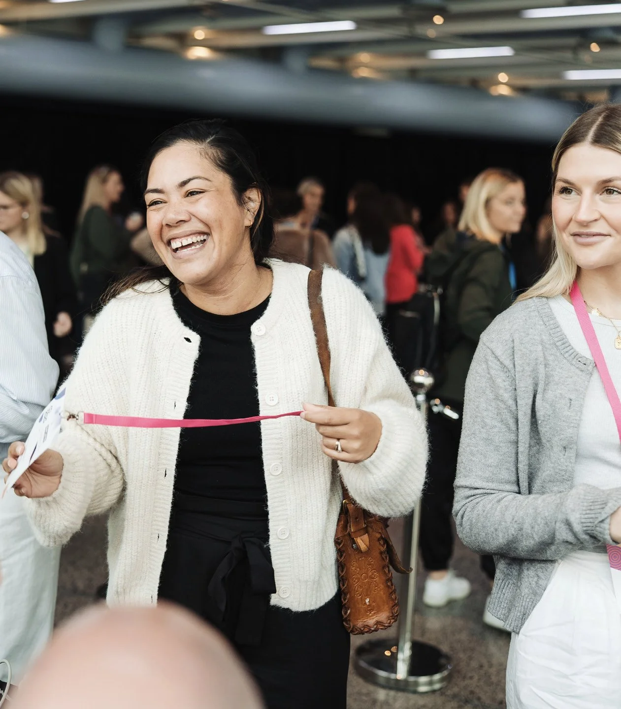 A group of people at an event, with a woman in the foreground smiling and holding a pink ribbon, and others in the background.