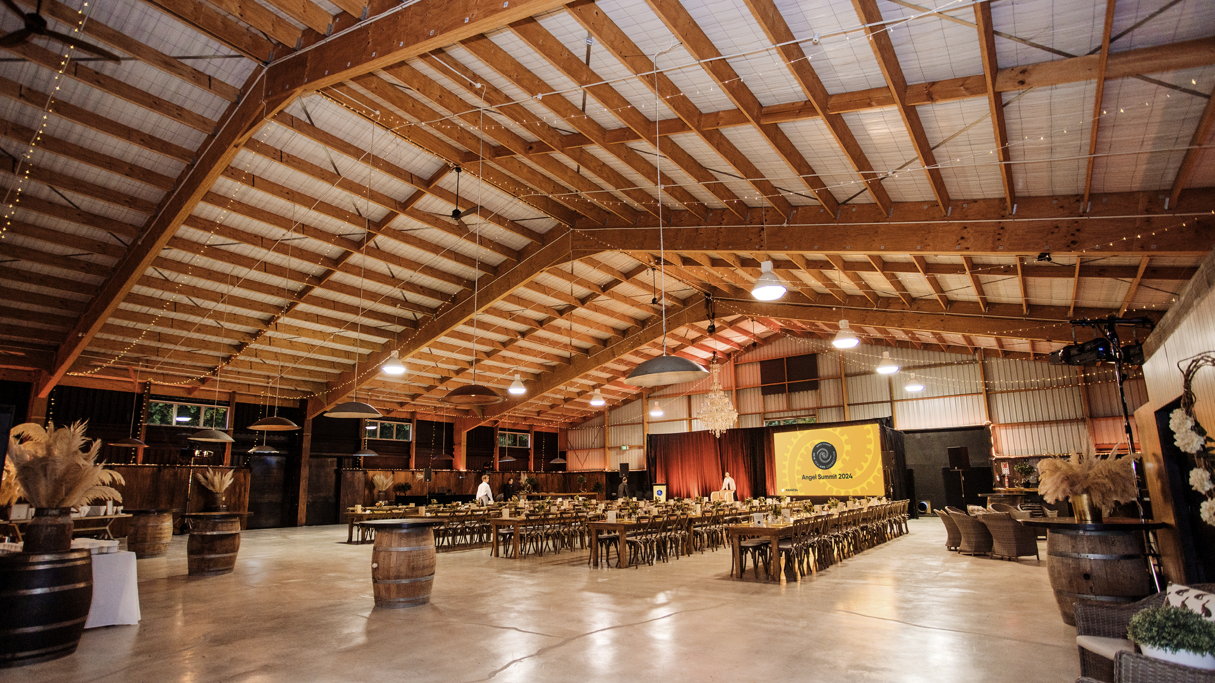 Interior of a large event hall with wooden beams, string lights, and tables arranged for a gathering, with a stage and a screen displaying 'Angel Summit 2024'.