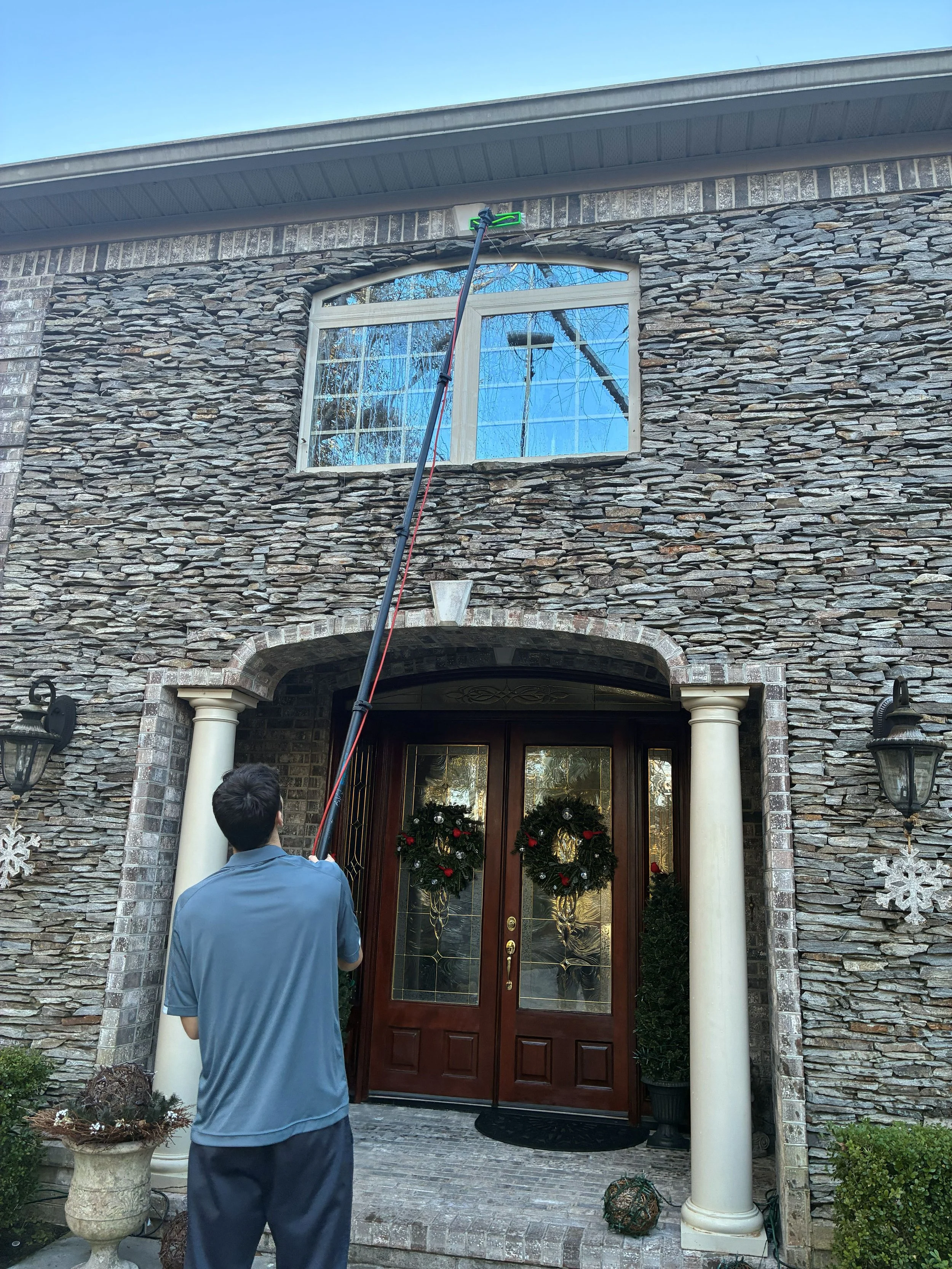 A man using a pole to clean the exterior window of a stone house decorated for Christmas with wreaths on the door and potted plants on each side.
