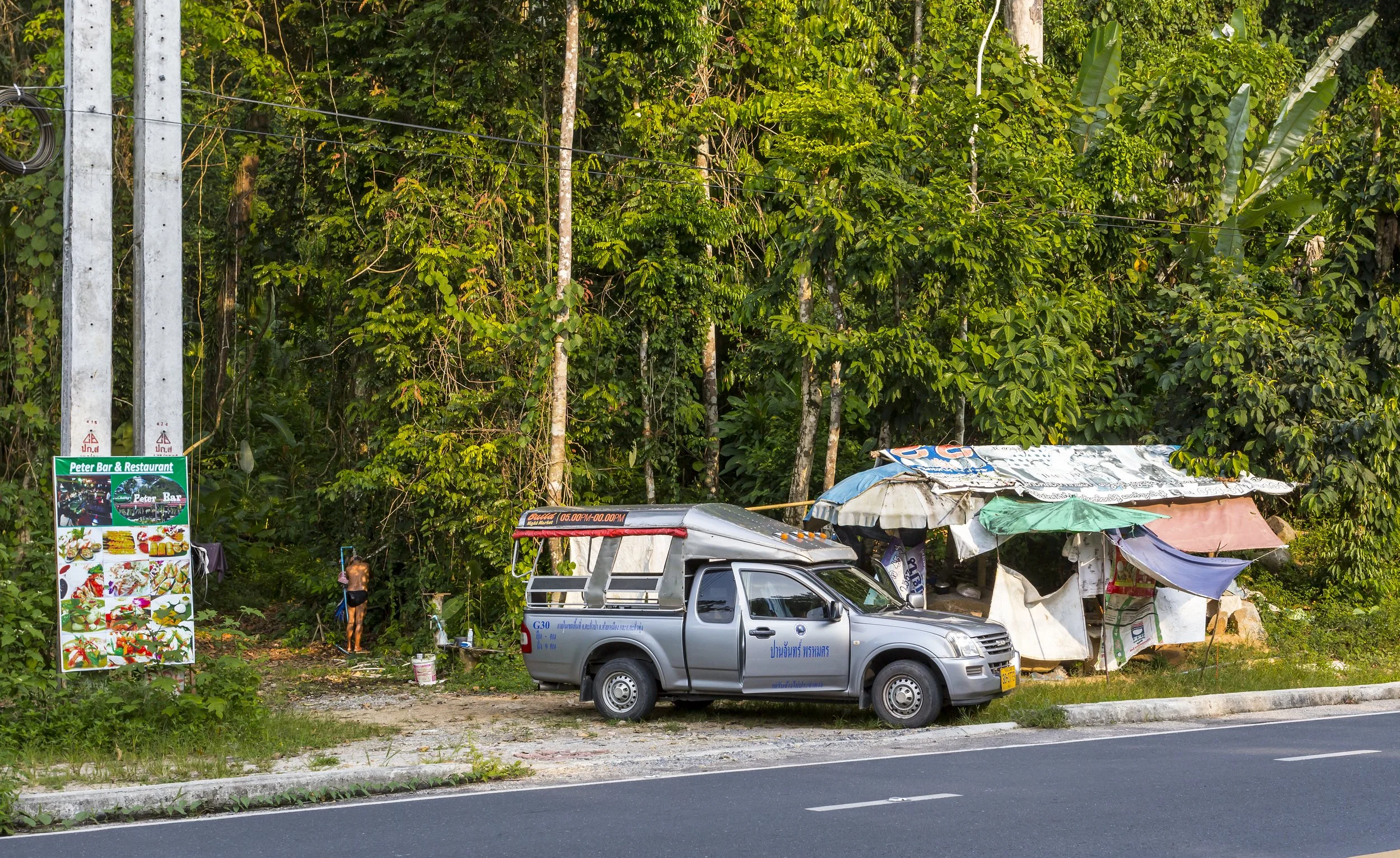 A roadside scene with a silver pickup truck parked on the side of a road near a makeshift shelter made of tarps, in front of a dense green forest.