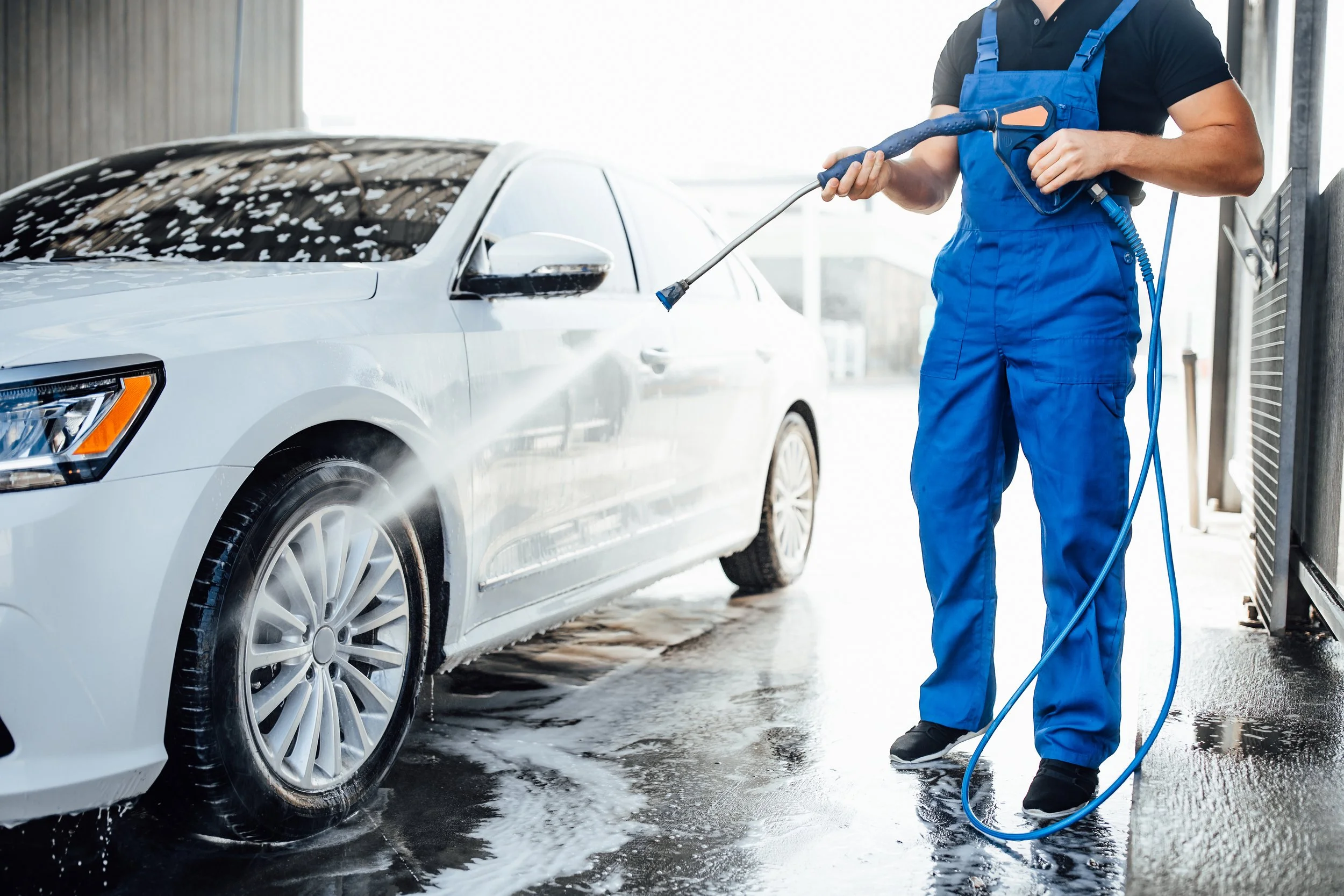 Person washing a white sedan with a pressure washer in a car wash