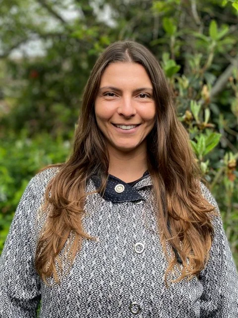 A woman with long brown hair smiling outdoors with greenery in the background.