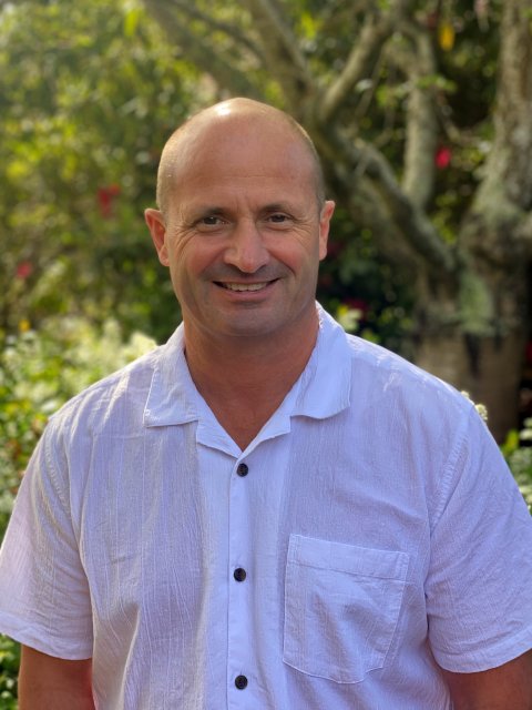 A smiling bald man wearing a white short-sleeve button-up shirt standing outdoors with trees and greenery in the background.