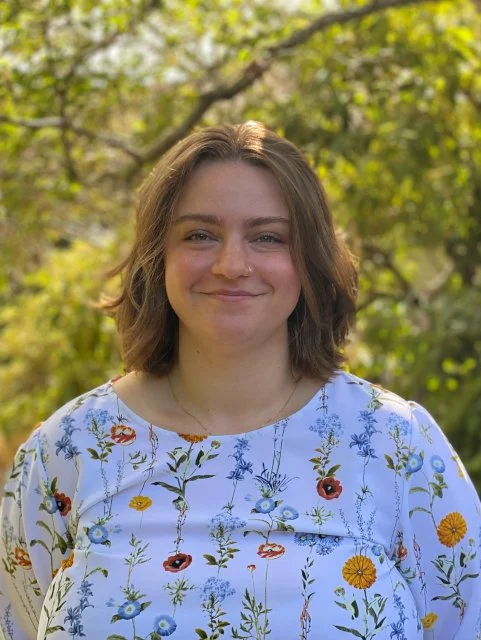 Young woman with shoulder-length brown hair smiling outdoors, wearing a white floral blouse, with trees and sunlight in the background.