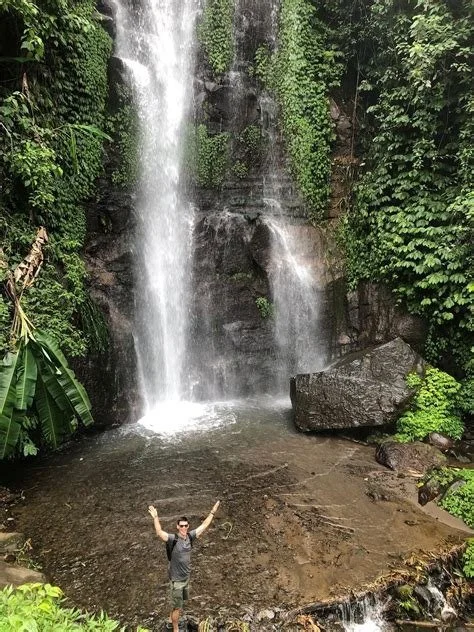 Man standing in front of a waterfall with arms raised, surrounded by lush green vegetation.