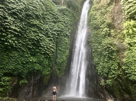 Person standing at the base of a tall waterfall surrounded by lush greenery.