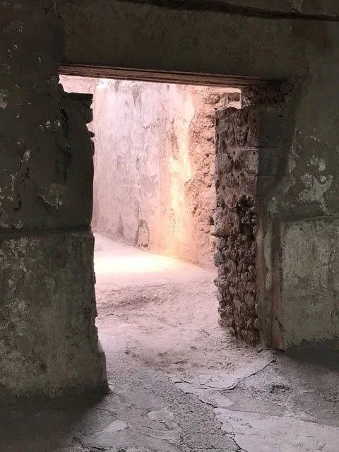 View of a narrow, rough stone passageway seen through a small, broken window opening in an old stone wall.