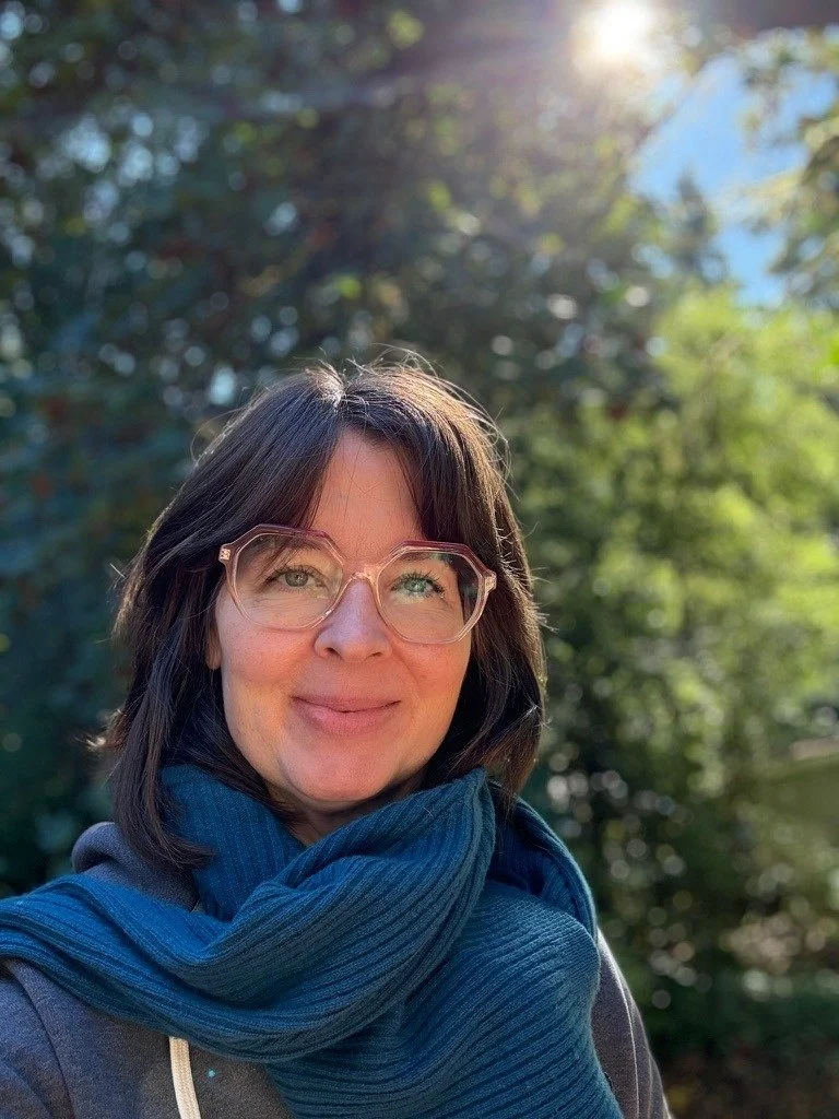 A woman with dark hair and glasses smiling outdoors with sunlight and green trees in the background.