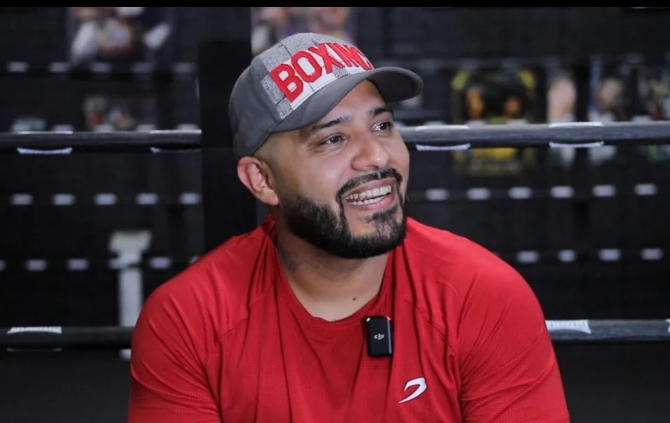 A man with a beard and mustache, wearing a red shirt and a gray baseball cap with red lettering, smiling and sitting in a boxing gym.