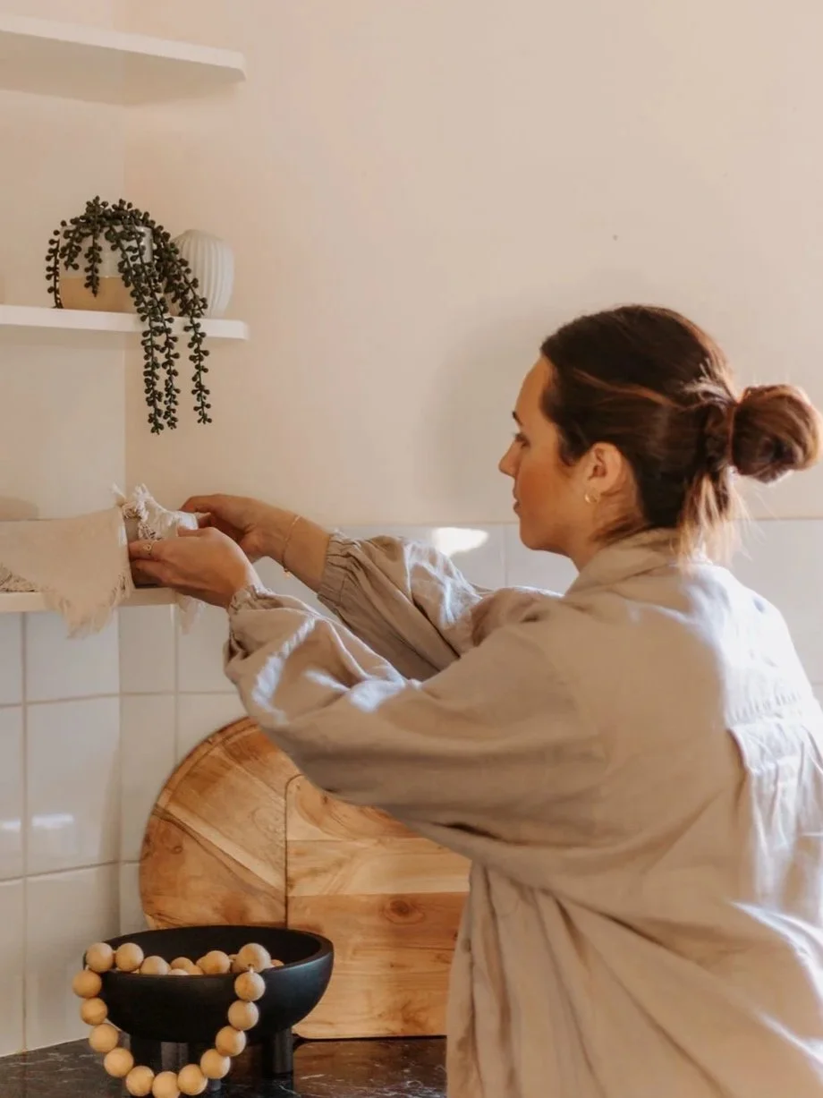 A woman with a bun hairstyle arranging items on a kitchen shelf, with a black bowl holding wooden beads and a wooden cutting board behind her.