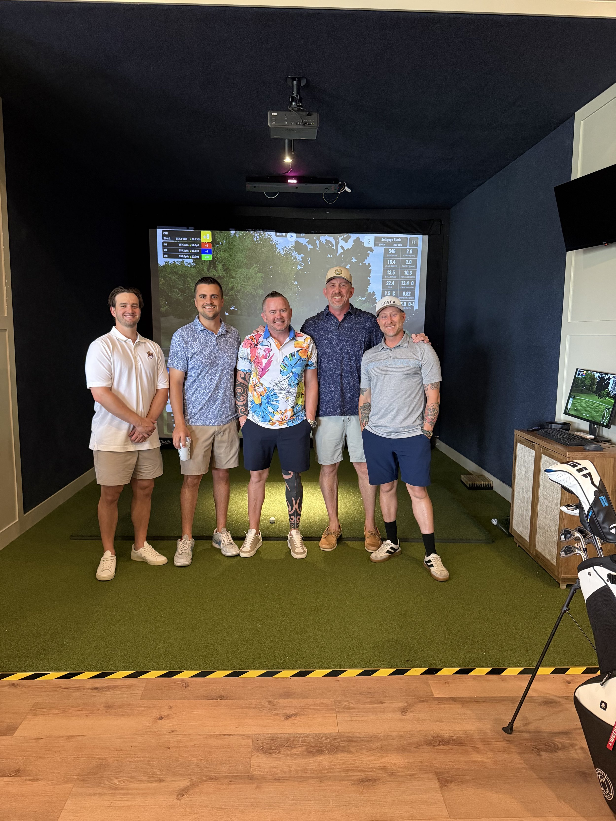 Five men standing side by side in an indoor golf simulator room, smiling at the camera. Behind them is a large screen displaying golf statistics and a golf course scene, with golf clubs and a golf ball in the foreground.