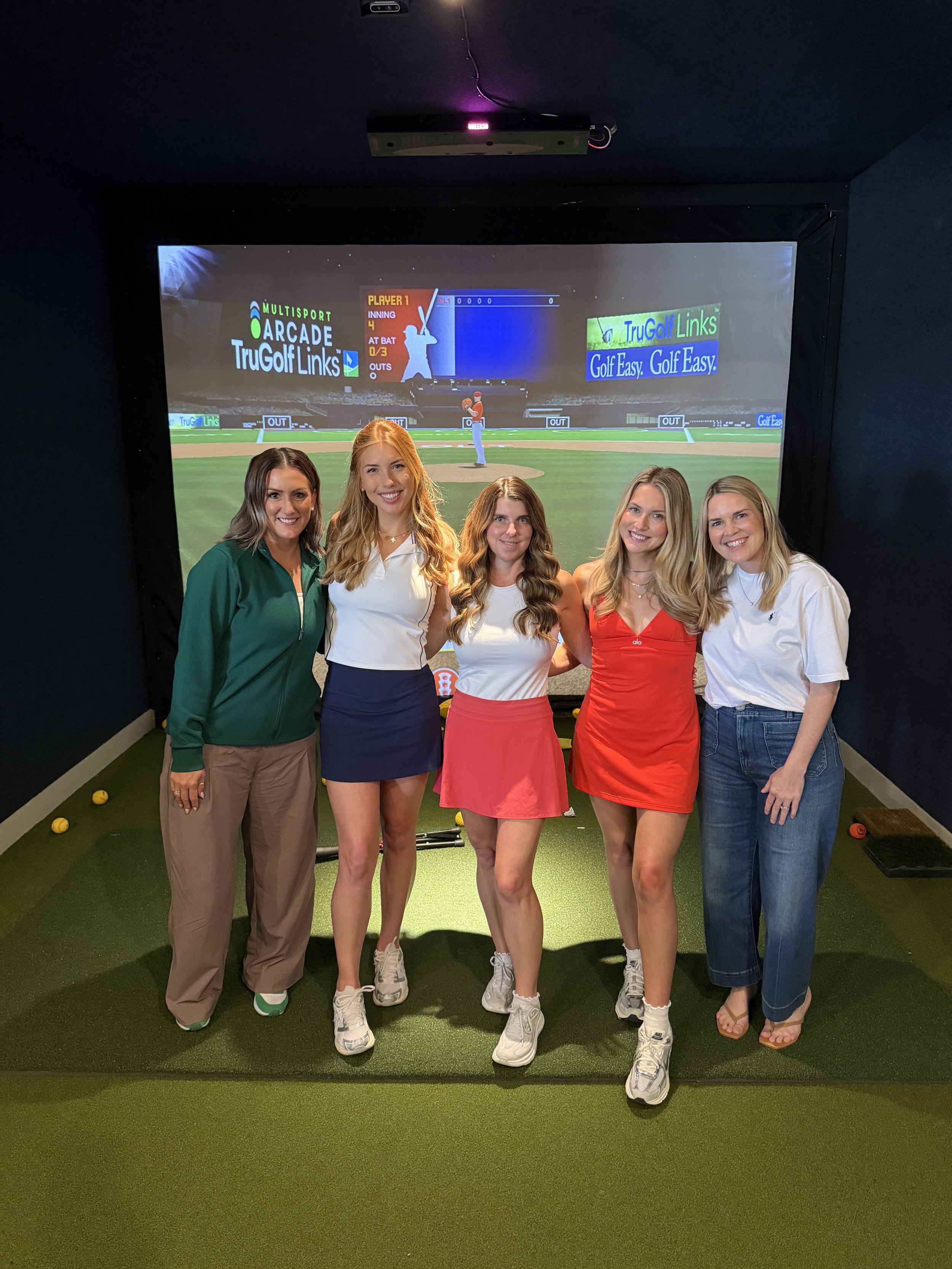 Five women standing together in an indoor golf simulation room, smiling at the camera. A large screen displaying a golf game is in the background.